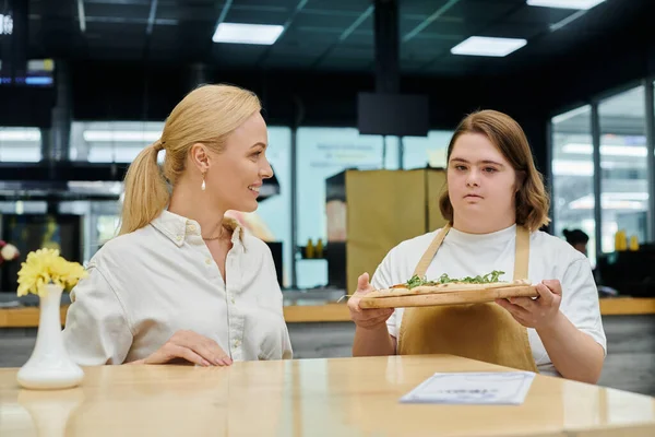 Young waitress with mental disability holding tasty pizza near smiling woman sitting in cafe — Stock Photo