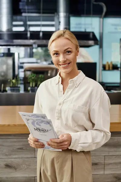Happy blonde administrator holding menu card and smiling at camera in modern cafe, small business — Stock Photo
