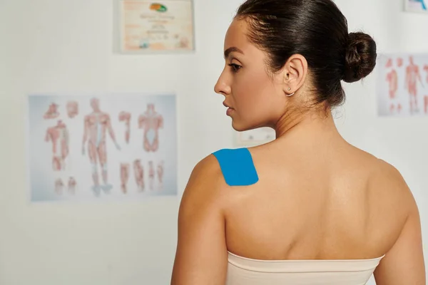 Vista posterior de la hermosa mujer joven con cintas de kinesio en el hombro durante la cita, atención médica - foto de stock