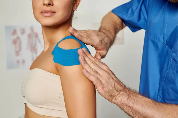 Vista ritagliata del medico mettere nastro kinesio sulla spalla della sua giovane paziente femminile, assistenza sanitaria — Foto stock