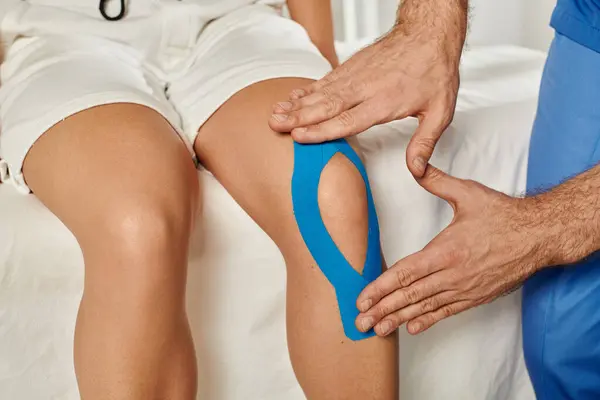 Vista recortada de la mujer en la cita mientras el médico pone cintas de kinesio en la rodilla, atención médica - foto de stock