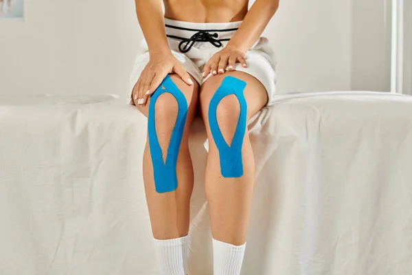 Visão cortada da jovem mulher sentada em sua enfermaria com fitas cinesiológicas nos joelhos, cuidados de saúde — Fotografia de Stock