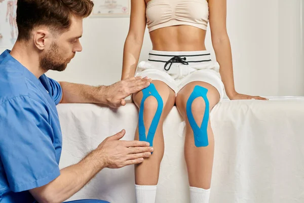 Bearded doctor putting kinesiological tapes on knee of his sitting female patient during appointment — Stock Photo