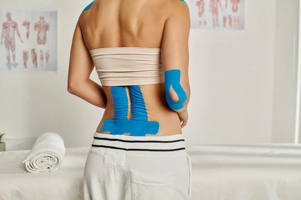 Vista recortada de una paciente joven de pie con cintas de kinesio en la espalda y el codo, atención médica - foto de stock