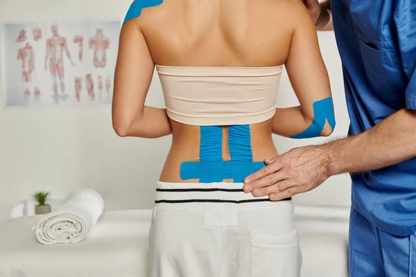 Vista posterior de la mujer en cita con el médico que pone cintas de kinesio en su cuerpo, atención médica - foto de stock