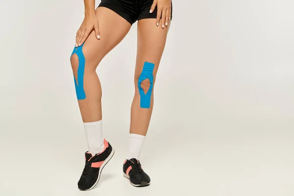 Visão cortada de pernas de mulher jovem com fitas cinesiológicas em seus joelhos no fundo cinza — Fotografia de Stock