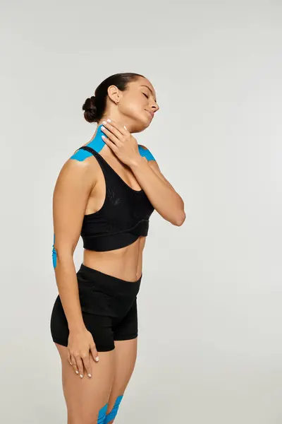 Bela mulher em traje esporte preto com fitas cinesiológicas em seus ombros tocando seu pescoço — Fotografia de Stock