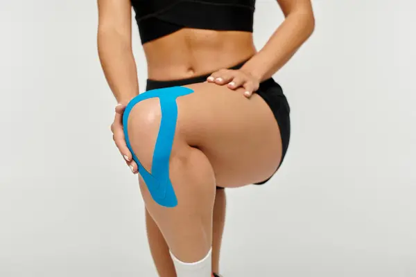 Vista ritagliata di giovane donna in costume sportivo in posa con nastri cinesiologici sul ginocchio, gamba sollevata — Foto stock