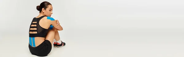 Belle jeune femme en tenue de sport assise sur le sol avec des bandes kinésiologiques sur le dos, bannière — Photo de stock