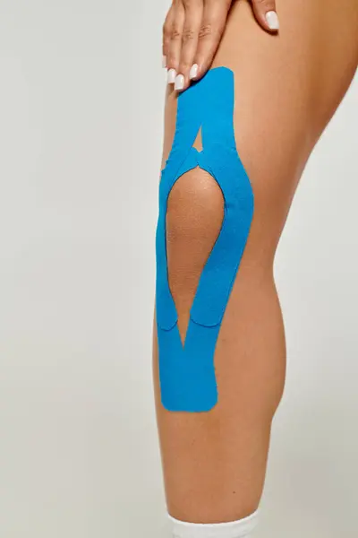 Vista recortada de mujer joven con cintas cinesiológicas azules en su rodilla posando sobre fondo gris - foto de stock
