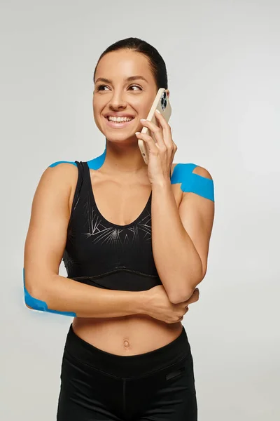 Mulher atraente alegre no desgaste do esporte com fitas kinesio no cotovelo e ombros falando por telefone — Fotografia de Stock