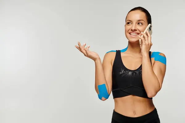 Веселая молодая женщина в черной спортивной одежде с кинезиологическими лентами, радостно разговаривающая по телефону — стоковое фото