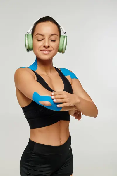 Привлекательная веселая женщина в спортивной одежде с наушниками и кинезиологическими лентами на локтях и плечах — стоковое фото