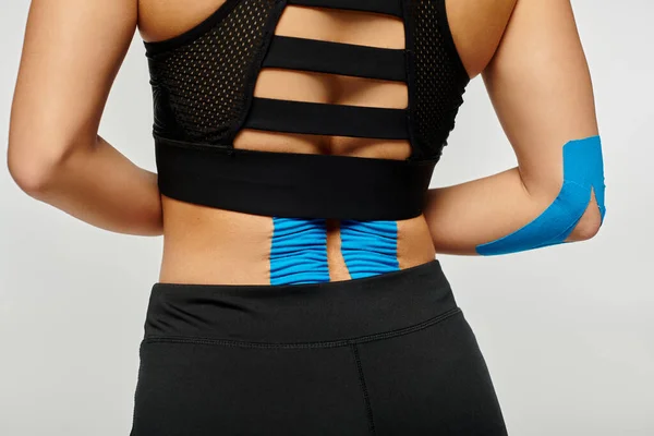 Vue recadrée de la jeune femme en tenue de sport avec des bandes kinésiologiques sur le dos sur fond gris — Photo de stock