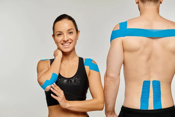 Cheerful woman in black sport attire smiling at camera next to sporty man with kinesiological tapes — Stock Photo