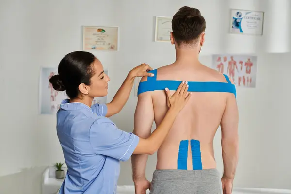 Beautiful young doctor in medical costume putting kinesio tapes on shoulders of her bearded patient — Stock Photo