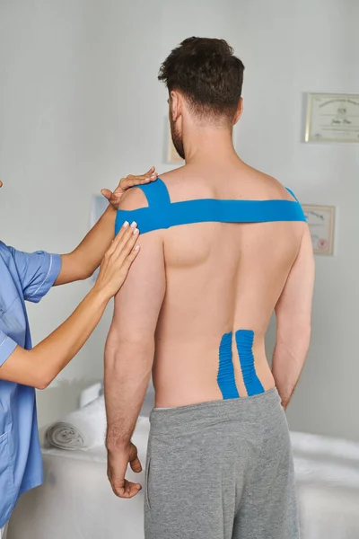Vista cortada de jovem médico ao lado de seu paciente masculino em calças de treino com fitas cinesiológicas — Fotografia de Stock