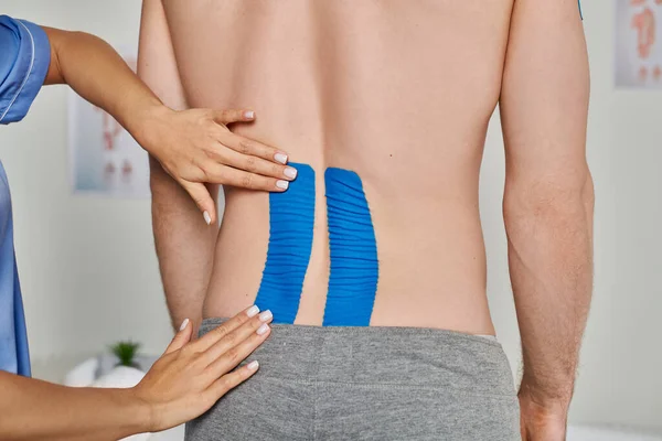 Cropped view of hands of female doctor putting kinesio tapes on back of her male patient, healthcare — Stock Photo