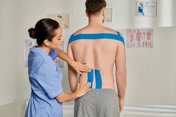 Attraktive junge Ärztin in medizinischem Kostüm legt ihrem Patienten kinesiologische Tapes auf den Rücken — Stockfoto