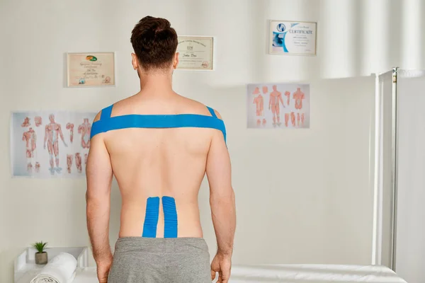 Visão traseira do paciente masculino em calças de suor cinza com fitas cinesiológicas em seu corpo, cuidados de saúde — Fotografia de Stock