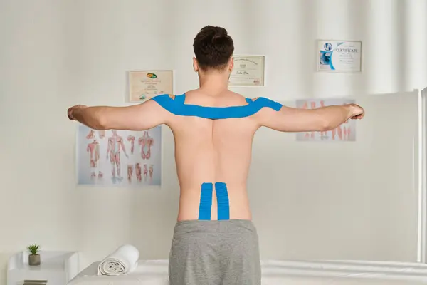 Visão traseira do paciente do sexo masculino posando com braços de lado com fitas cinesiológicas em seu corpo, cuidados de saúde — Fotografia de Stock