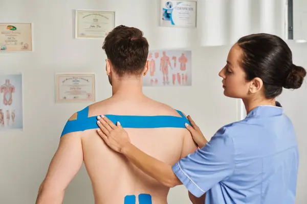 Красивий молодий лікар кладе кінезіологічні стрічки на спину і плечі свого пацієнта, охорона здоров'я — стокове фото