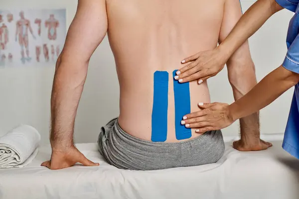 Vista recortada de la joven doctora poniendo cintas de kinesio azul en la espalda de su paciente, cuidado de la salud - foto de stock