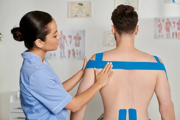 Attraktive Ärztin in blauem Medizinkostüm, die Kinesiobänder auf den Rücken ihrer Patientin legt, Gesundheitswesen — Stockfoto