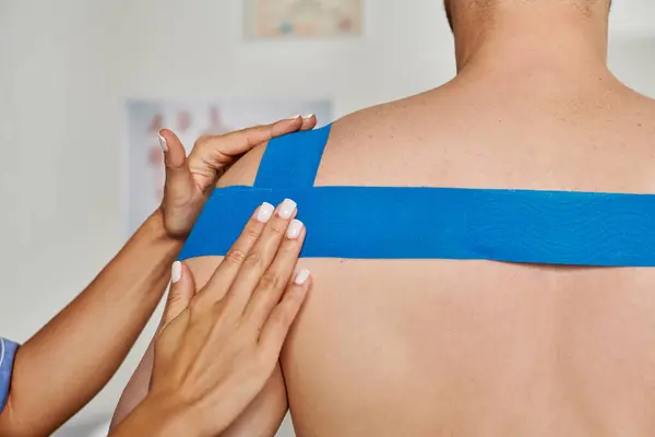 Vista ritagliata della mano del giovane medico femminile che mette il nastro kinesio sulla spalla del suo paziente — Foto stock