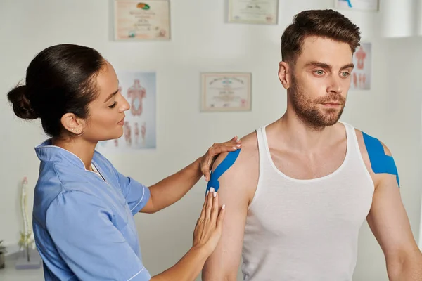 Attractive young doctor in blue medical costume putting kinesiological tapes on her handsome patient — Stock Photo
