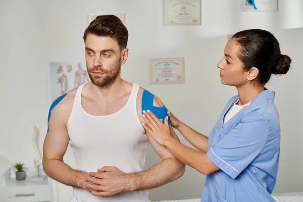 Attractive young doctor in medical costume looking at her handsome patient with kinesio tapes — Stock Photo