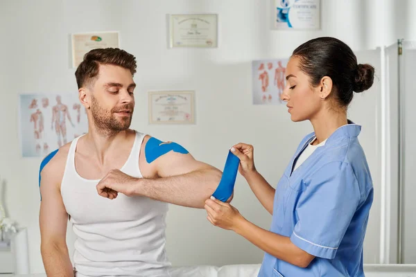 Jolly handsome patient sitting and watching his doctor putting kinesiological tape on his elbow — Stock Photo