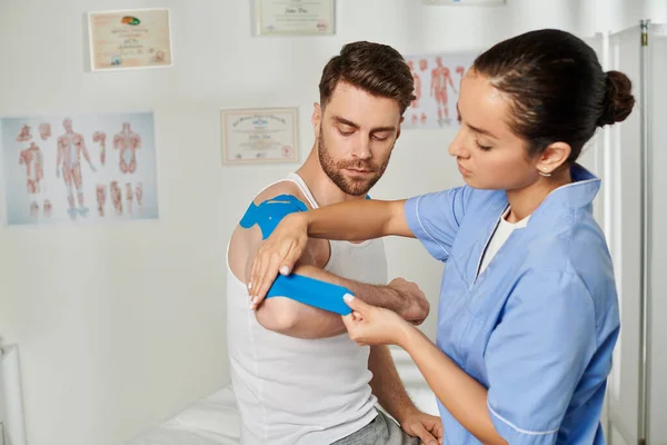 Good looking man sitting and watching his young doctor putting kinesio tape on his elbow, healthcare — Stock Photo