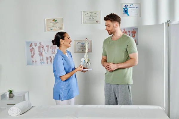 Веселий лікар, який показує модель хребта своєму привабливому пацієнту під час призначення, охорона здоров'я — стокове фото