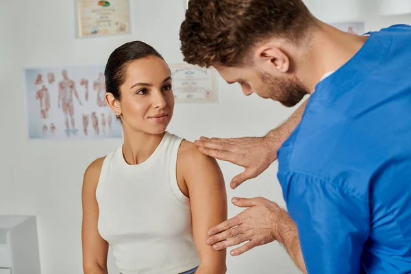 Beautiful young woman looking at her bearded doctor while he checking her shoulder, healthcare — Stock Photo