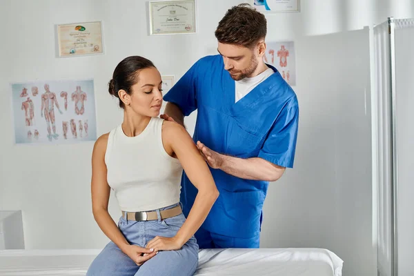 Handsome doctor in blue medical costume massaging shoulder of his young attractive patient — Stock Photo