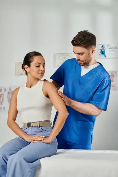 Приваблива молода жінка в повсякденному вбранні дивиться, як лікар перевіряє своє плече, охорона здоров'я — стокове фото