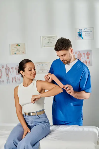 Mujer joven con atuendo casual viendo cómo el médico pone cintas de kinesio en su codo, atención médica - foto de stock