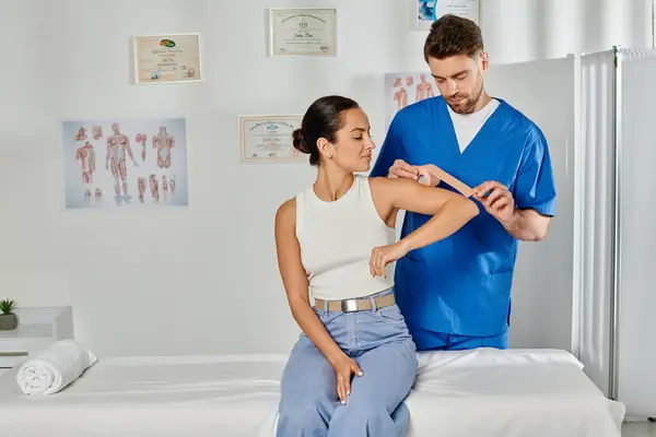 Attractive young woman in casual attire looking at her doctor putting kinesio tape on her elbow — Stock Photo