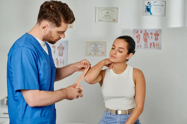 Gut aussehender Arzt in medizinischem Kostüm legt kinesiologische Klebebänder auf Körper seiner Patientin — Stockfoto
