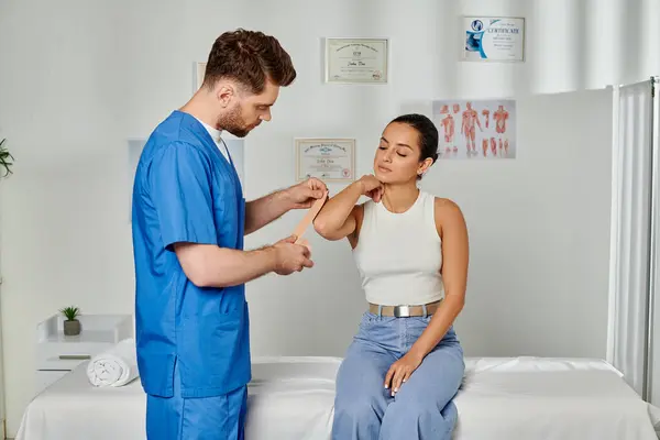 Guapo doctor barbudo poniendo cinta cinesiológica en el codo de su joven paciente, cuidado de la salud - foto de stock