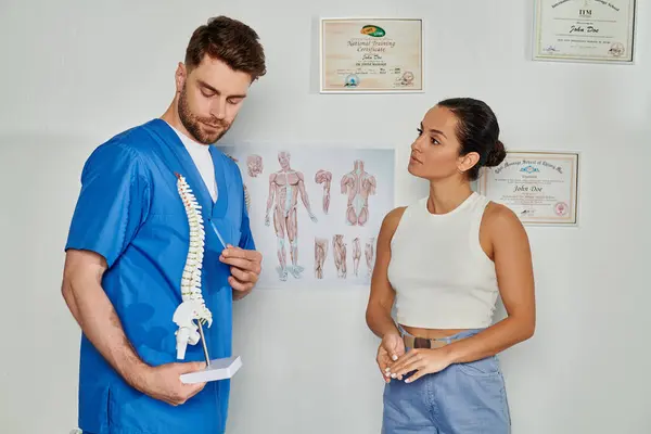Médico bonito em azul traje médico mostrando modelo da coluna vertebral para seu jovem paciente do sexo feminino, cuidados de saúde — Fotografia de Stock