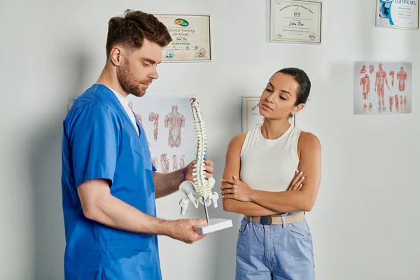 Atractivo mujer paciente mirando ner guapo barbudo médico con columna vertebral modelo en sus manos - foto de stock