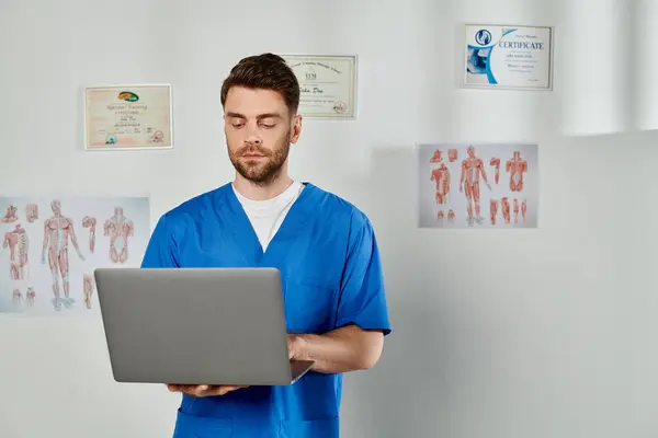 Gut aussehender bärtiger Arzt in blauer medizinischer Tracht arbeitet an seinem Laptop, Gesundheitswesen — Stockfoto