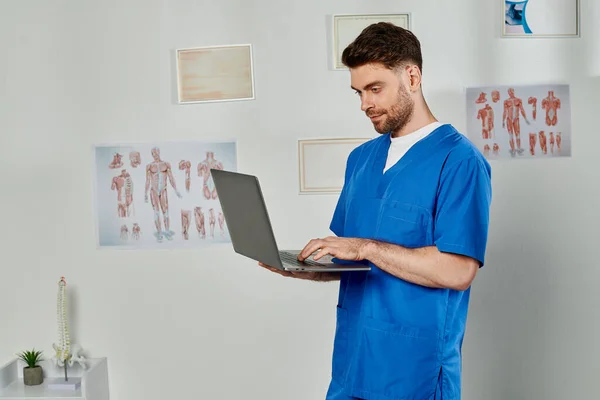 Bom olhar médico barbudo olhando para seu laptop enquanto trabalhava duro em seu escritório, cuidados de saúde — Fotografia de Stock