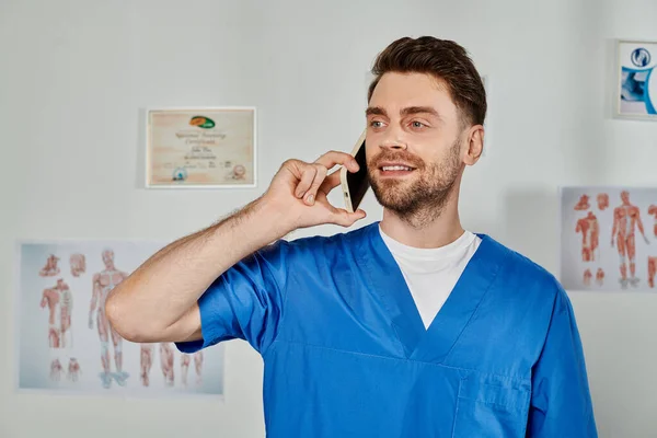 Alegre bonito barbudo médico consultoria por telefone e sorrindo enquanto em seu escritório, cuidados de saúde — Fotografia de Stock