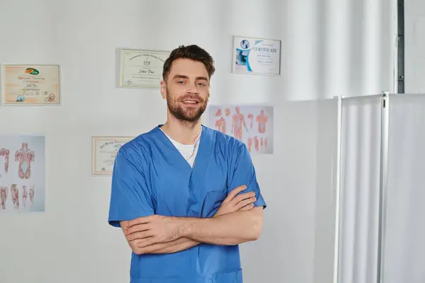 Alegre médico guapo posando con los brazos cruzados en el pecho y sonriendo a la cámara, atención médica - foto de stock