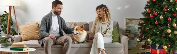 Happy couple sitting on couch and cuddling corgi dog near Christmas tree and presents, banner — Stock Photo