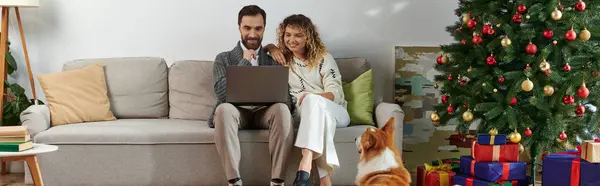 Banner, man using laptop and sitting on couch with curly wife near cute corgi dog and Christmas tree — Stock Photo