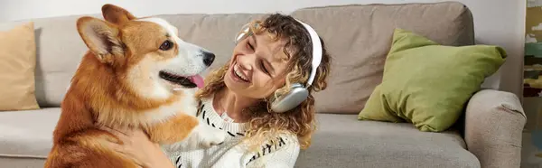 Curly woman in wireless headphones playing with cute corgi dog in modern apartment, happy moments — Stock Photo